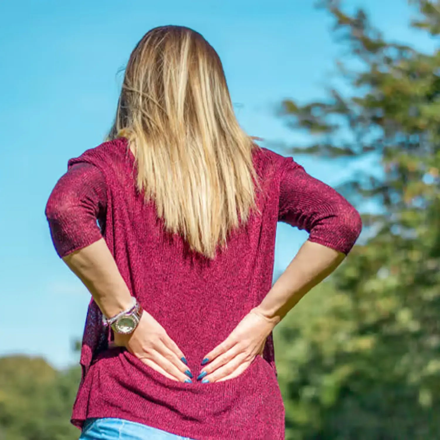 A woman with long, light brown hair, wearing a maroon sweater and blue jeans, stands outdoors with her hands on her lower back, indicating pain.